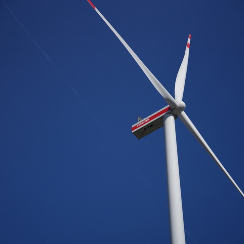 Nahaufnahme einer Windturbinenschaufel vor blauem Himmel, mit sichtbarem Turm und Rotorbl&auml;ttern.