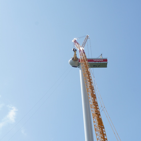 Blick nach oben auf einen Windkraftanlagen-Mast mit Baustellenkran und Arbeitsplattform im blauen Himmel.