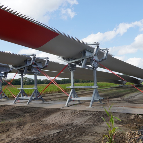 Nahaufnahme von Windradfl&uuml;geln, die auf speziellen Halterungen in einem Feld fixiert sind. Wolken am Himmel.