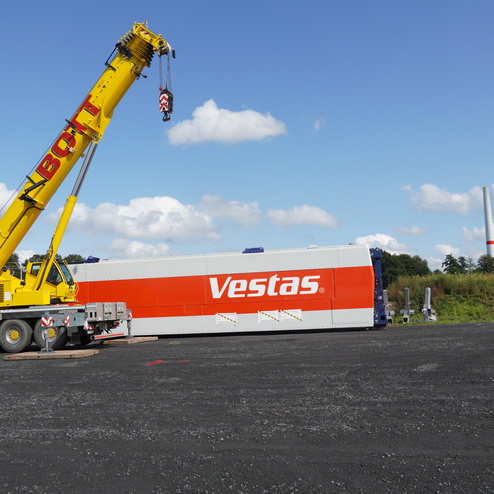 Gelber Kran hebt ein Vestas-Windradteil auf einer Baustelle mit grauem Untergrund und blauem Himmel im Hintergrund.