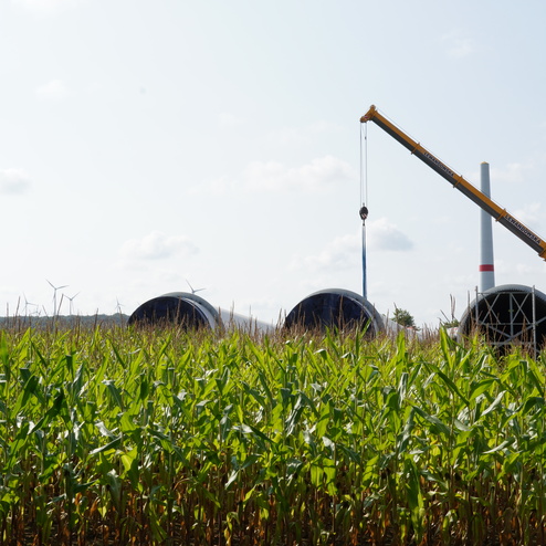 Kran hebt gro&szlig;e Bauteile &uuml;ber ein Feld mit Maispflanzen; im Hintergrund sind Windkraftanlagen sichtbar.