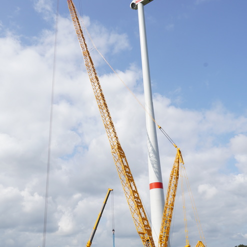 Gro&szlig;er Kran hebt Rotorbl&auml;tter zu einer Windkraftanlage unter einem bew&ouml;lkten Himmel auf einem gr&uuml;nen Feld.