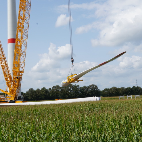 Gabelstapler hebt ein Windradblatt &uuml;ber ein Feld, w&auml;hrend ein Kran im Hintergrund am Windkraftwerk arbeitet.
