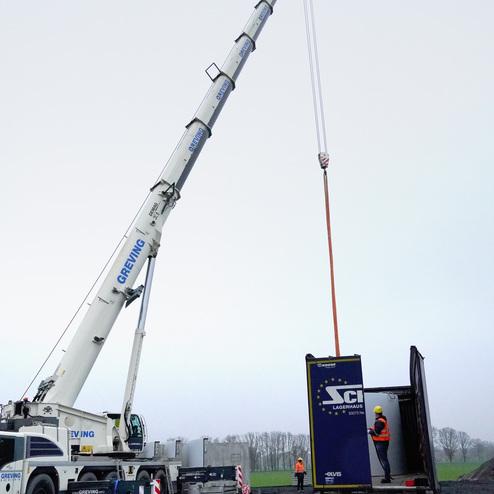 Kran hebt einen Container von einem Lkw auf einer Baustelle, Arbeiter stehen in Sicherheitskleidung in der N&auml;he.