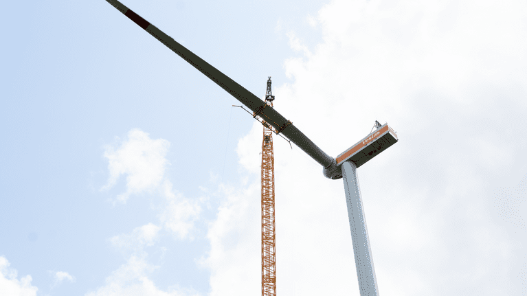 Blick auf einen Windkraftanlagenrotor und einen Kran vor einem wolkenverhangenen Himmel.