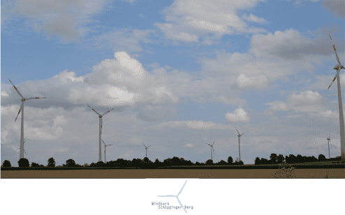 Windkraftanlagen stehen vor einem bew&ouml;lkten Himmel, umgeben von landwirtschaftlichen Fl&auml;chen und B&auml;umen.