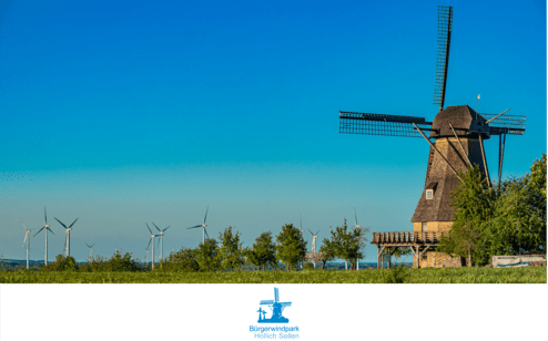 Windm&uuml;hle neben Windkraftanlagen vor blauem Himmel, umgeben von B&auml;umen und Wiese im B&uuml;rgerwindpark Hollich Seilen.