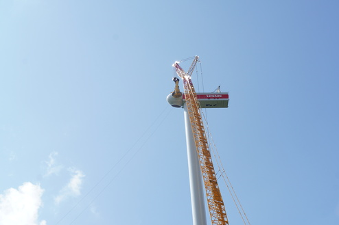 Blick nach oben auf einen Kran, der eine Windkraftanlage mit orangefarbenem Turm und blauem Himmel in der N&auml;he hebt.