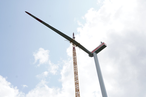 Blick auf eine Windkraftanlage mit einem Kran, der das Rotorblatt anbringt, vor einem bew&ouml;lkten Himmel.