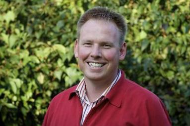 L&auml;chelnder Mann mit kurzen Haaren, rot-wei&szlig; gestreiftem Hemd und rotem Pullover vor gr&uuml;ner Pflanzenwand.