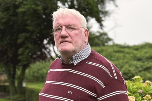 &Auml;lterer Mann in gestreiftem Pullover steht im Garten vor Str&auml;uchern und dekorativen Schafsskulpturen.