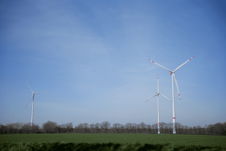 Drei Windkraftanlagen stehen auf einem gr&uuml;nen Feld unter einem blauen Himmel mit wenigen Wolken.