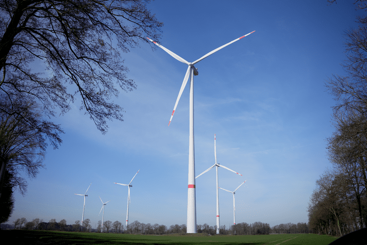 Windkraftanlagen auf einem Feld unter blauem Himmel, umgeben von B&auml;umen und Wiesen.